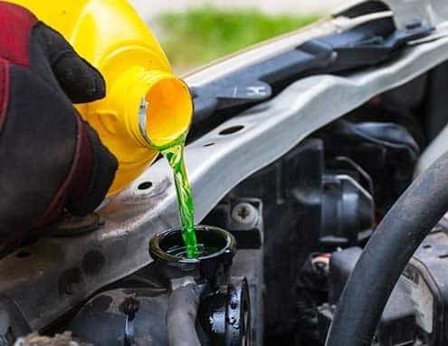 A Mechanic wearing red gloves is pouring bright green engine coolant into a car radiator under the bonnet to keep the cooling system at a safe operating temperature.