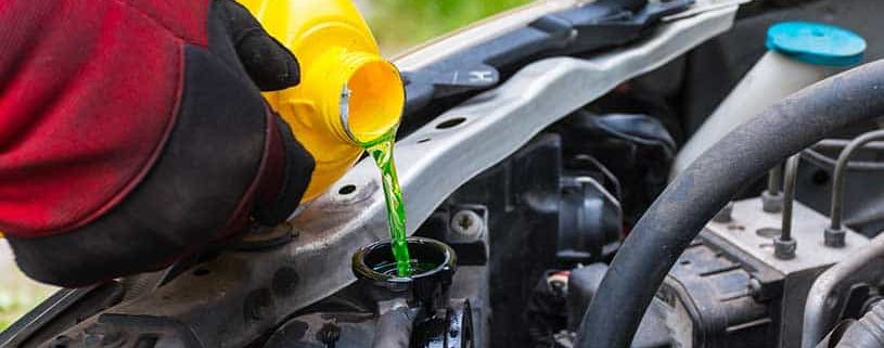 A Mechanic wearing red gloves is pouring bright green engine coolant into a car radiator under the bonnet to keep the cooling system at a safe operating temperature.