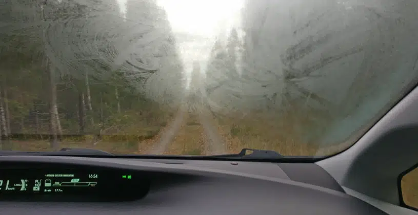A view from inside a car showing a heavily fogged and smeared windscreen, obscuring the dirt road and trees ahead.