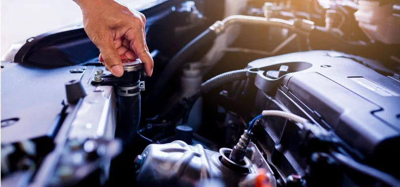 A hand twists a radiator cap inside a car engine bay, showing hoses and mechanical parts under the open bonnet.