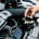 A hand removes the radiator cap from a car engine bay, showing various mechanical components under the open bonnet.