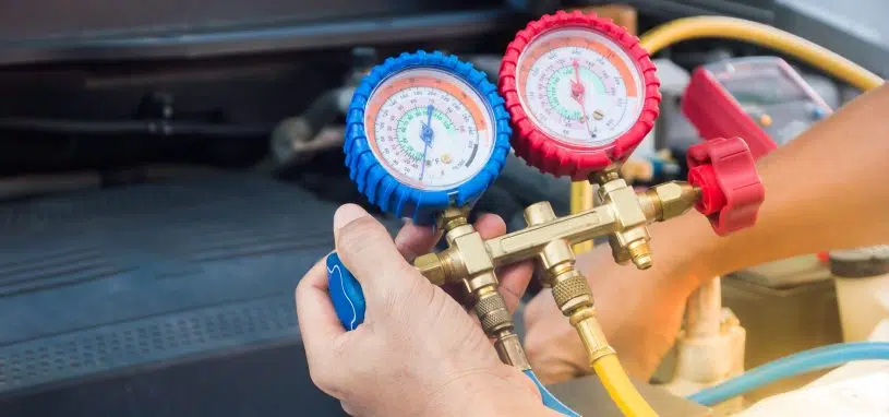 Hands hold a manifold gauge set with blue and red dials to check a car's air conditioning system over an open engine bay.