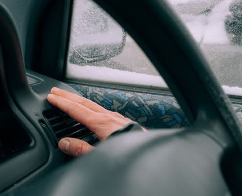 A hand hovers over a car dashboard vent to check the heater airflow, seen through a steering wheel on a snowy day.