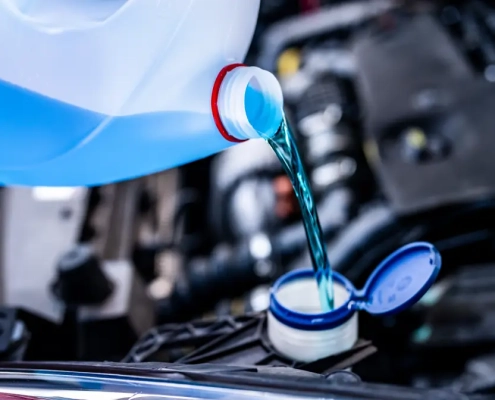 Green engine coolant being poured from a white container into a car’s coolant reservoir under the hood.
