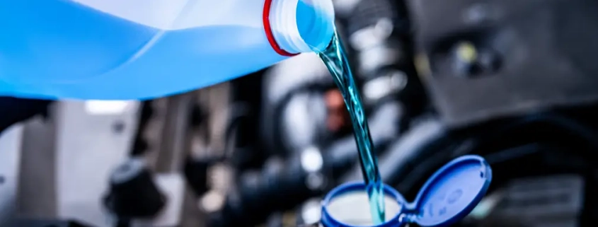 Green engine coolant being poured from a white container into a car’s coolant reservoir under the hood.