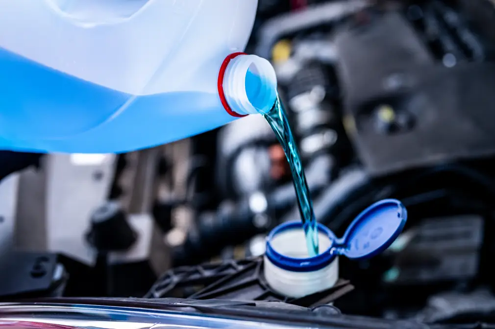 Green engine coolant being poured from a white container into a car’s coolant reservoir under the hood.