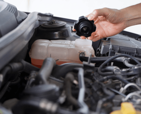 Close-up of a car engine with a hand opening the coolant reservoir cap, showing the cooling system components under the bonnet.