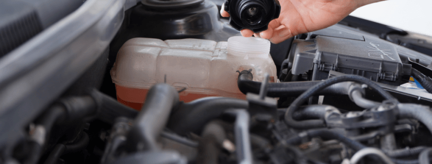 Close-up of a car engine with a hand opening the coolant reservoir cap, showing the cooling system components under the bonnet.