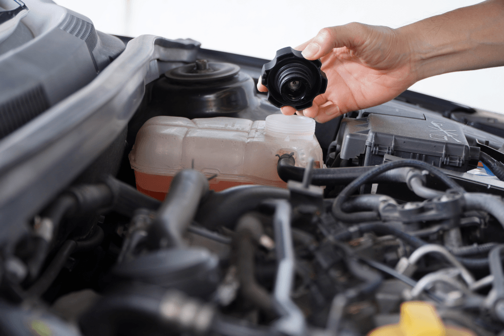 Close-up of a car engine with a hand opening the coolant reservoir cap, showing the cooling system components under the bonnet.