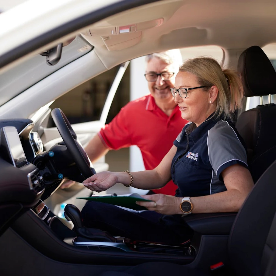 A female technician in a Natrad uniform sits in a car, discussing a clipboard with a smiling customer in a red shirt.
