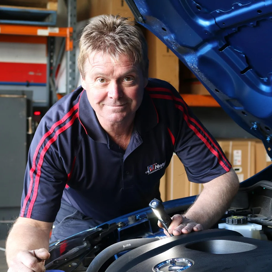 A smiling mechanic in a navy and red polo shirt uses a screwdriver while working on a blue car’s engine in a workshop.