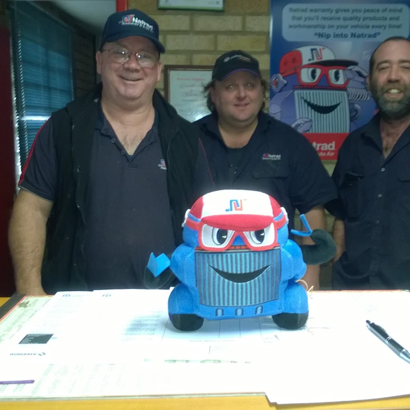 Three smiling Natrad staff members stand behind a counter, featuring a blue cartoon radiator mascot plush.