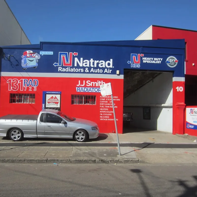 A blue and red Natrad workshop with JJ Smith signage, a silver ute parked in front, and a large open garage bay door.