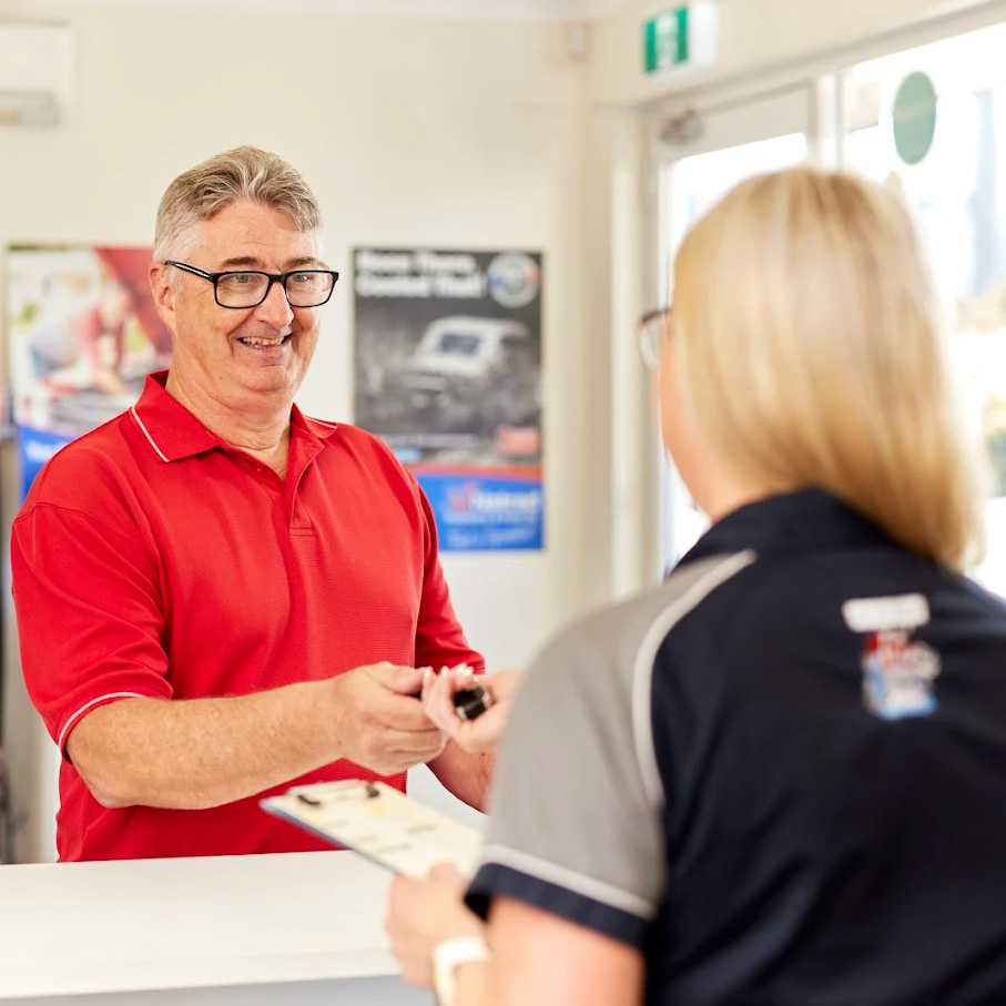 A smiling customer in a red shirt hands car keys to a Natrad staff member at a service counter in a bright office.