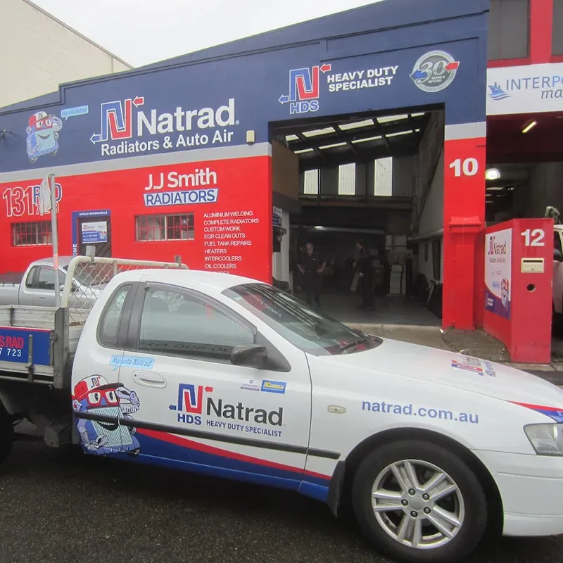 A white Natrad branded utility vehicle parked in front of the red and blue JJ Smith Radiators workshop with an open garage.